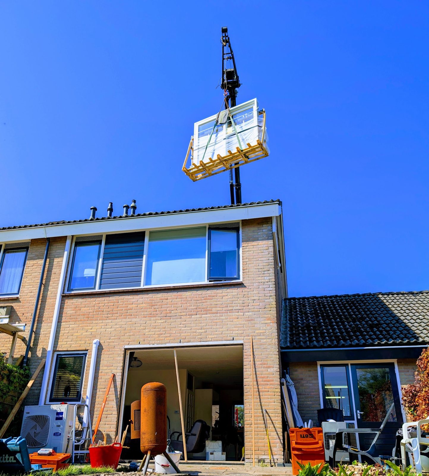 Een kraan hijst een cabine boven het dak van een woning, de garage staat open
